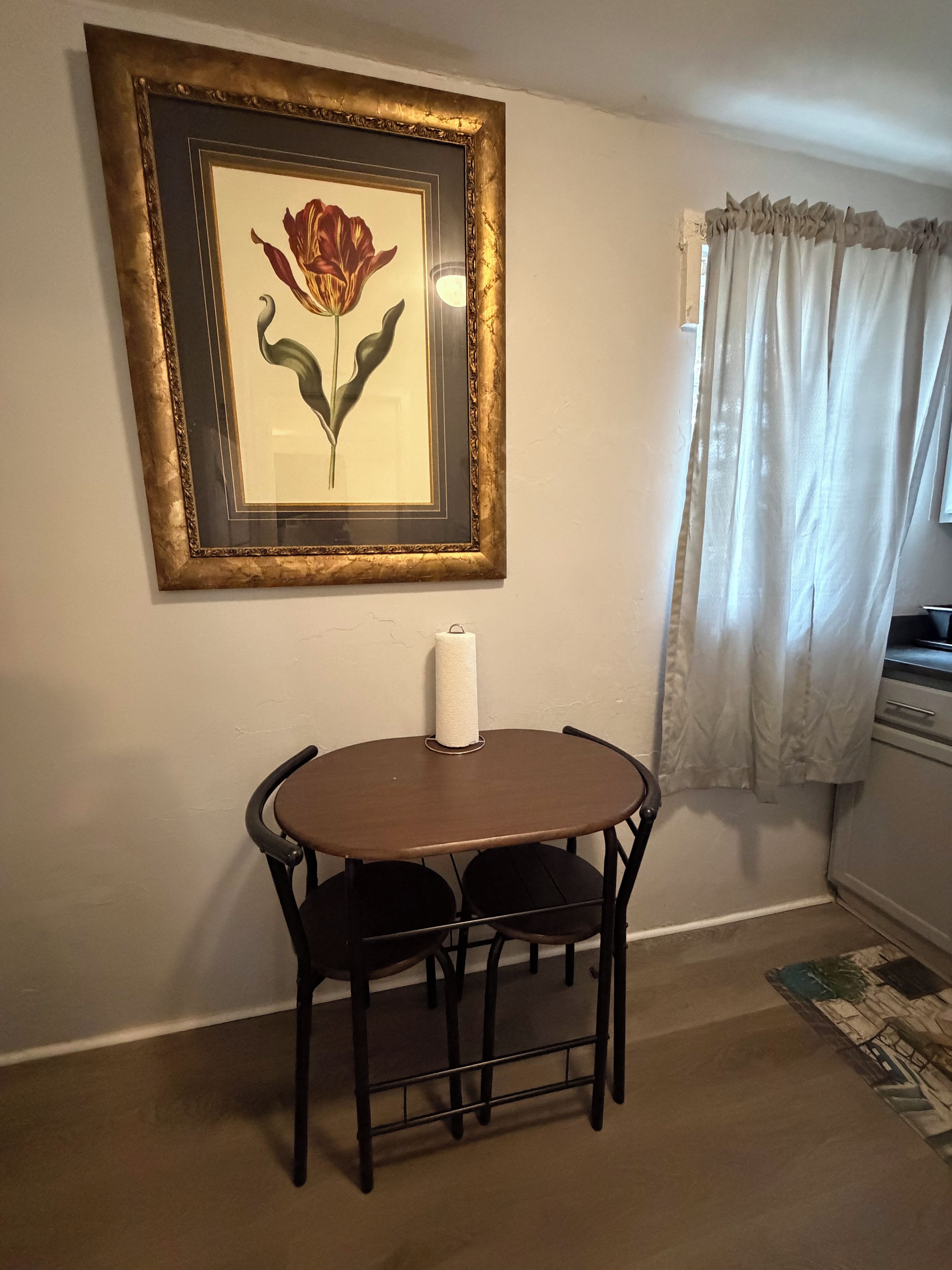A dining area with a table and chairs, a framed picture on the wall, and a window with curtains.