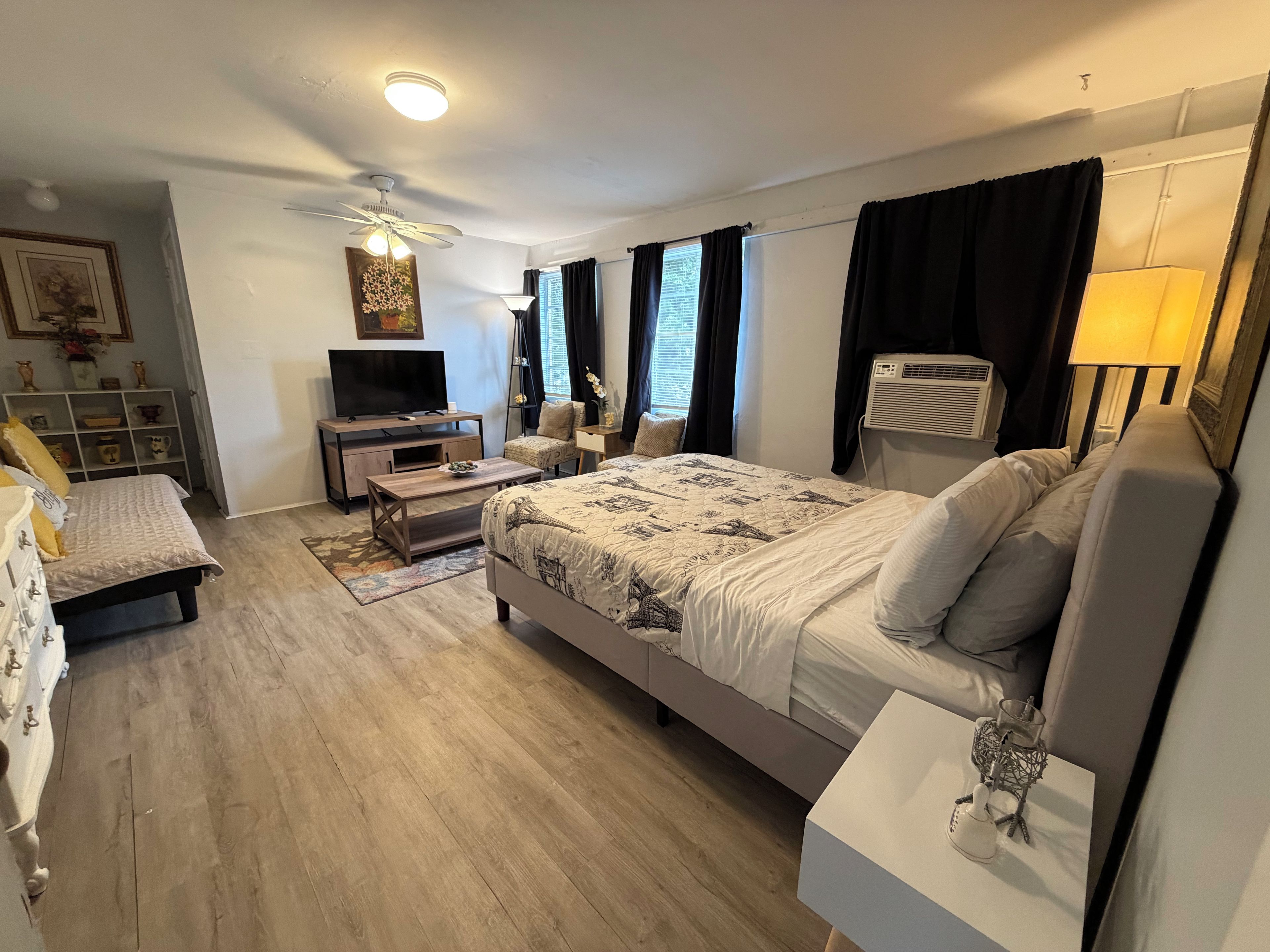 Une chambre à coucher comprenant un lit, des tables de chevet, un téléviseur, un ventilateur de plafond et une fenêtre avec des rideaux.