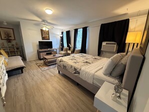 Un dormitorio con cama, mesitas de noche, televisión, ventilador de techo y ventana con cortinas.
