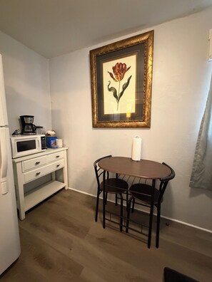 Eine kleine Küchenzeile mit einer Mikrowelle, einer Kaffeemaschine und Aufbewahrungsbehältern. Über einem Tisch mit zwei Stühlen hängt ein gerahmtes Blumenbild.