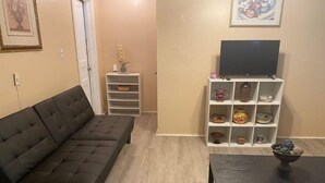 Uma sala de estar com um sofá marrom escuro, uma mesa pequena com uma tigela, uma TV em um móvel e uma estante branca com diversos objetos.