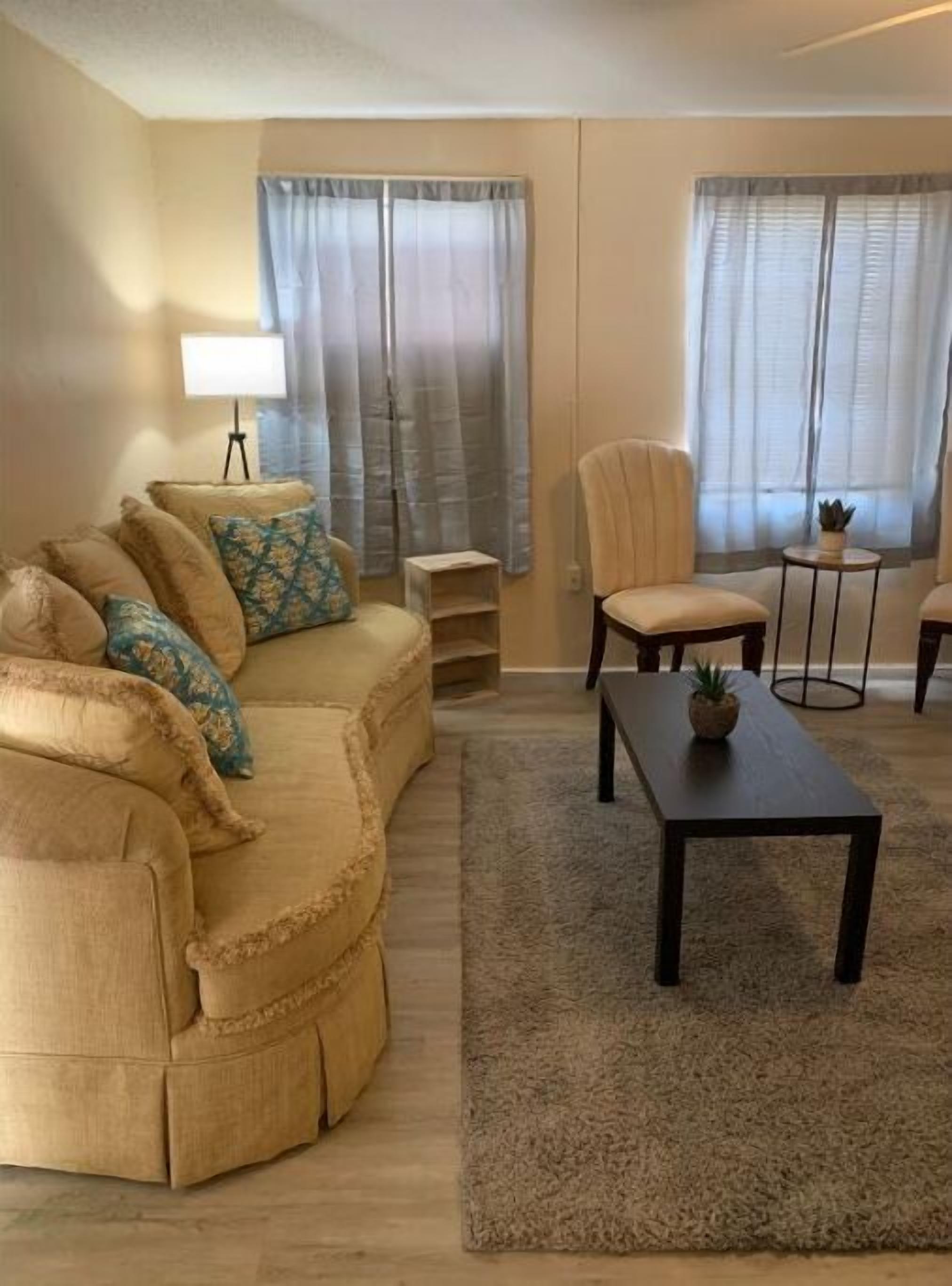 A living room with a beige sofa, a small round table, and a chair. There are two windows with sheer curtains.