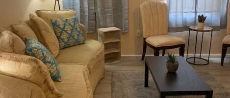 A living room with a beige sofa, a small round table, and a chair. There are two windows with sheer curtains.