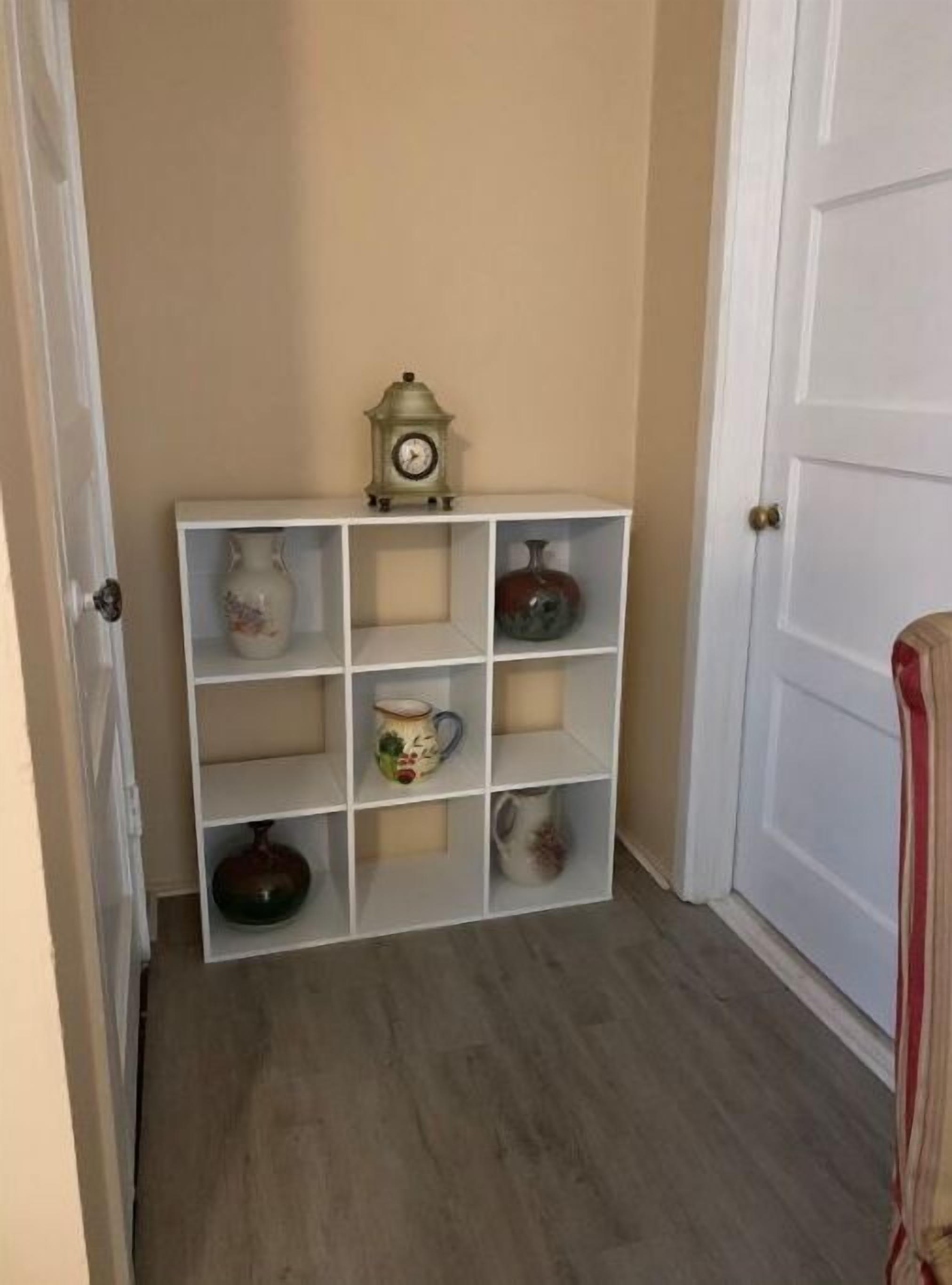 En hall med en vit hylla längs en beige vägg, där olika föremål av keramik och en klocka är placerade.