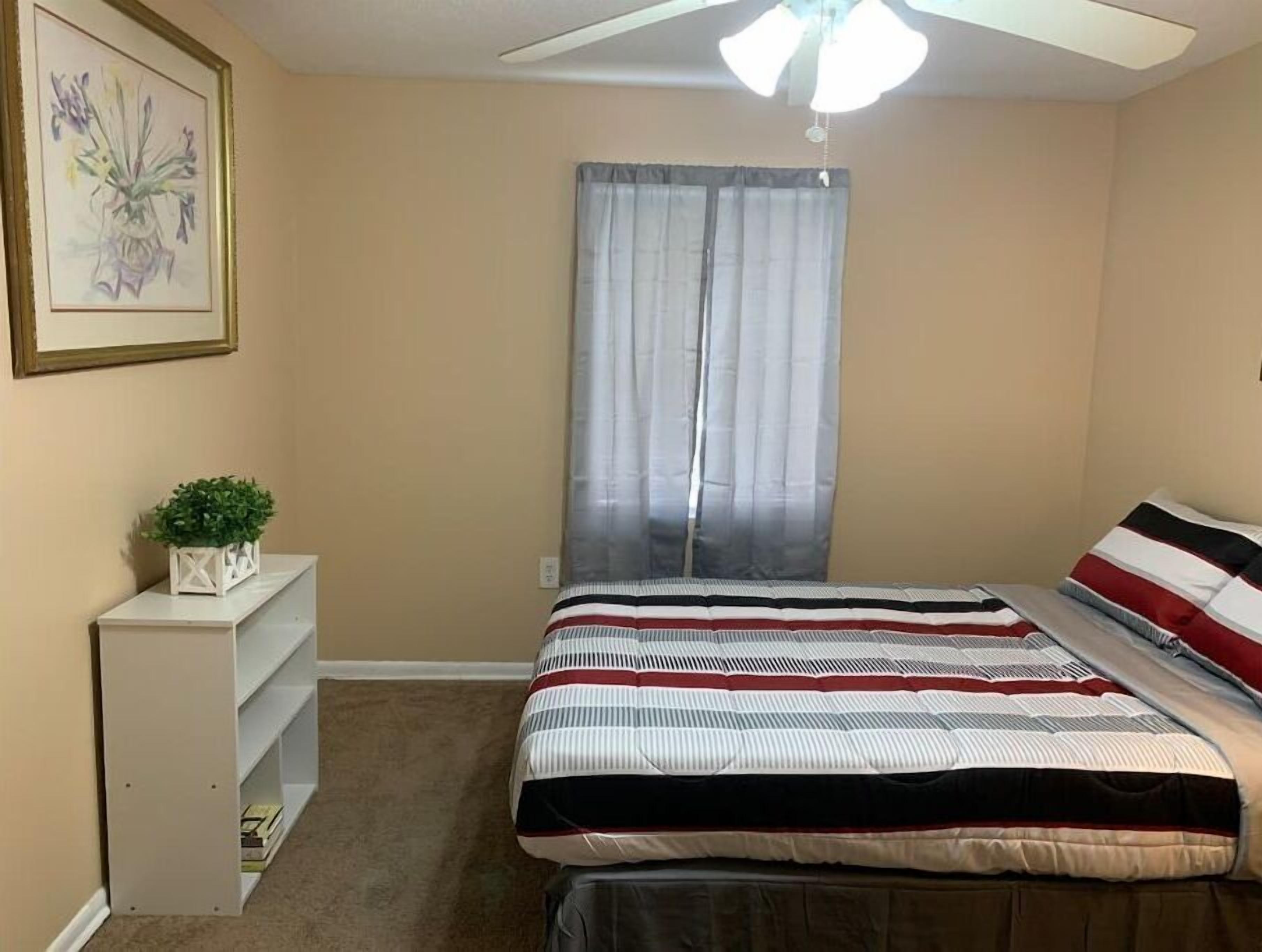 A bedroom with a bed, a ceiling fan, a window with curtains, a small plant on a side table, and a framed picture on the wall.