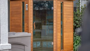 Una sauna de madera en esquina con puerta de vidrio y un pequeño espacio para sentarse al aire libre.