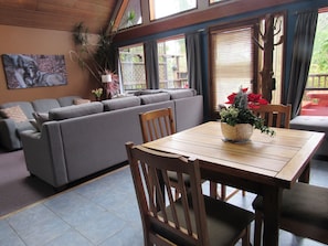 Una sala de estar con un sofá gris, una mesa de comedor de madera y sillas. Hay plantas en macetas sobre la mesa y un cuadro en la pared.