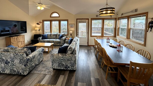 A spacious living room with a large sectional sofa, a dining table with chairs, a television mounted on the wall, and large windows providing natural light. - OS733- 6 Bedrooms- Tiki Bar- Private Pool and Hot Tub! (Corolla)
