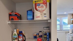 Una cocina con armarios y encimera blancos. Se observan varios utensilios de cocina en los estantes, incluyendo una caja de toallas de papel, una botella de detergente para platos y un tarro de mantequilla de cacahuete.
