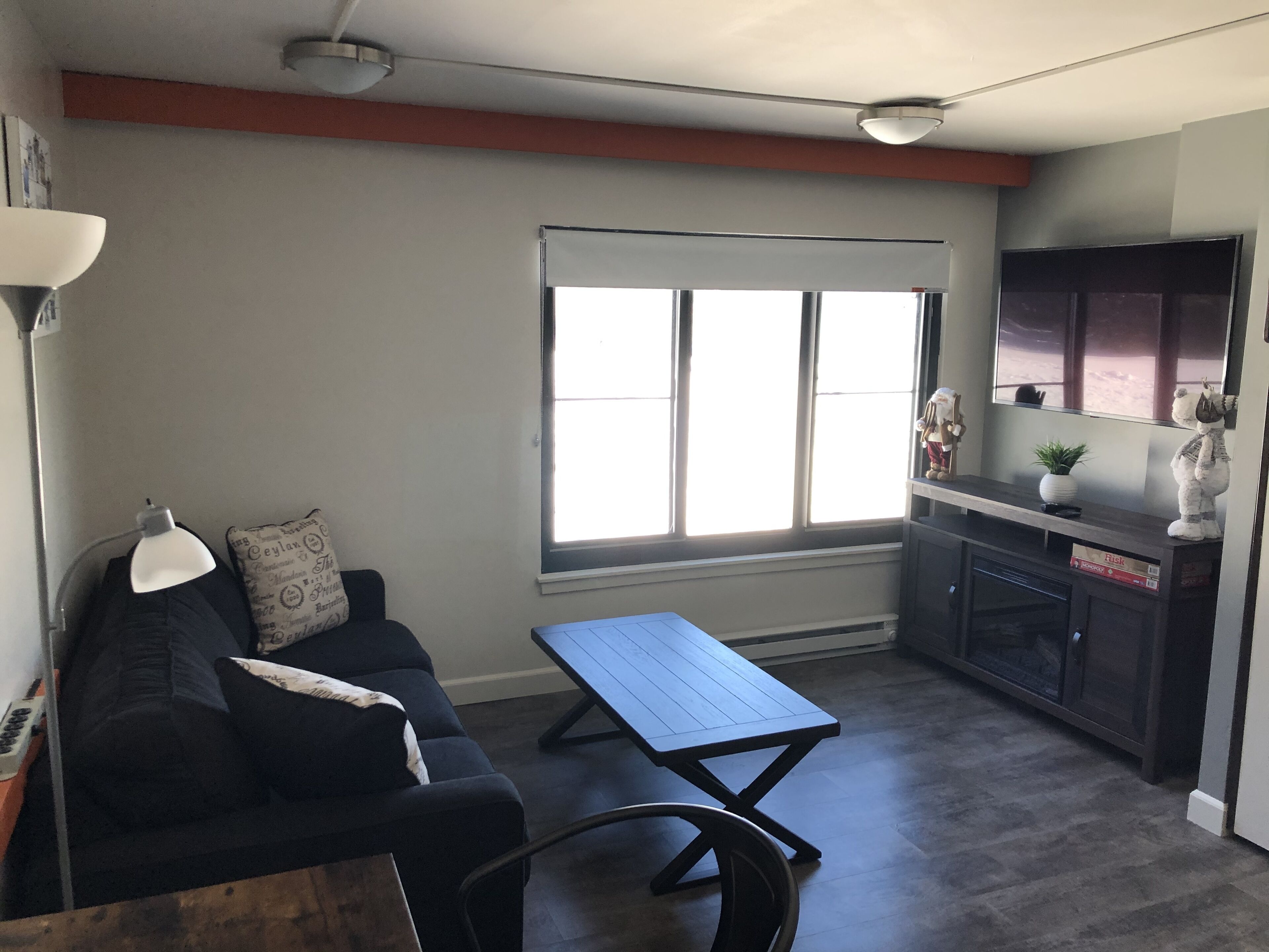 A compact living space with a sofa, a small table, a window with blinds, a kitchenette, and a ceiling light fixture.