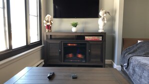 A living room with a fireplace, a television mounted on the wall, a sofa, and a coffee table with two remote controls. - SLOPES VIEW, SKI IN AND OUT AT SILVER CREEK, SLEEPS 4. (Snowshoe)