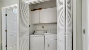 Une buanderie équipée de deux machines à laver et d’un sèche-linge, avec une étagère en bois au-dessus et une porte blanche dotée d’une poignée.