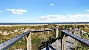 Une promenade en bois qui mène à une plage bordée de dunes, sous un ciel dégagé.