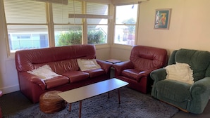 A living room with a red leather sofa, a green armchair, a small table, and a large window with blinds. - Perfect as a Falls Creek base camp. Pet friendly.  (Mount Beauty)