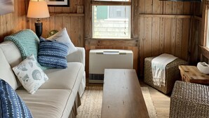 Un accogliente interno di una baita con soffitto in legno, un divano con cuscini decorati, un tavolo in legno, un televisore montato a parete e una finestra con persiane.