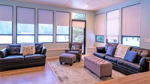 Ein geräumiges Wohnzimmer mit zwei Sofas, einem Sessel und einem Couchtisch. Große Fenster mit Jalousien lassen viel Tageslicht herein.