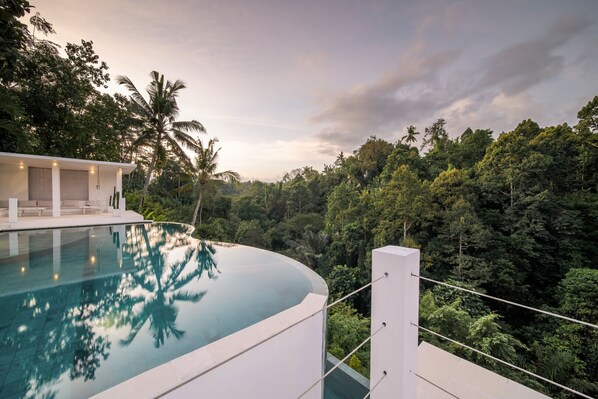 Une piscine à débordement moderne offrant une vue dégagée sur une végétation luxuriante et un bâtiment blanc doté d’un espace extérieur aménagé pour s’asseoir.