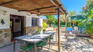 An outdoor patio with a table and chairs, a covered area with a thatched roof, and a pool in the background. - Casa Rural la Sacri Guaro by Ruralidays (Málaga)