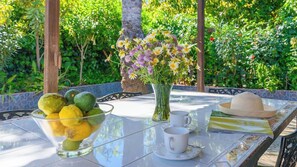 A table with a glass bowl of citrus fruits, a vase with flowers, white cups, and a straw hat.
