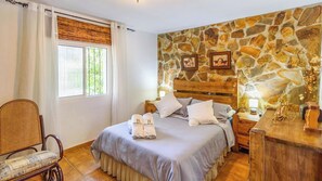 A bedroom with a stone wall, a bed with pillows, a chair, and a nightstand with a lamp.
