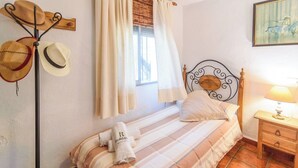 A bed with striped bedding, a bedside table with a lamp, and a wall-mounted hat rack with several hats.