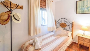 A bed with striped bedding, a bedside table with a lamp, and a wall-mounted hat rack with several hats.