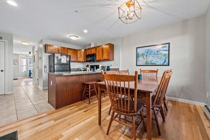 A modern kitchen with wooden cabinets, a central island, and a dining area with wooden chairs. - Mountain Brook 152 - Ski in Ski out at Saddleback (Sandy River Plantation)