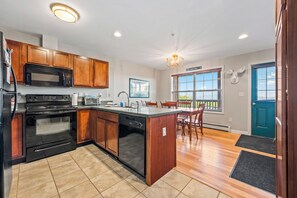 A modern kitchen with wooden cabinets, a built-in oven, microwave, and dishwasher, a large island with seating, and a view of the outdoors through a window. - Mountain Brook 152 - Ski in Ski out at Saddleback (Sandy River Plantation)