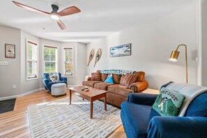 A modern living room with a ceiling fan, a brown leather sofa, a blue armchair, and a coffee table. - Mountain Brook 152 - Ski in Ski out at Saddleback (Sandy River Plantation)