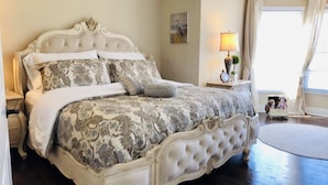 Een klassiek hemelbed met een bloemenbedekking en passende kussens, een nachtkastje met een lamp, en een klein tafeltje met beeldjes.