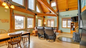 Amplia sala de estar con mesa de comedor de madera, dos sillones de cuero, chimenea y ventanas grandes que ofrecen vistas al exterior.