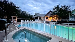 Una piscina con jacuzzi, rodeada por una cerca blanca, y un edificio con un porche al fondo.