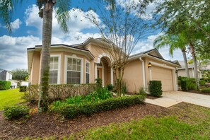 A residential house with a beige exterior, a garage, and a well-maintained garden with various plants and a palm tree. - Beautiful Pool Home in Gated Community Close to Disney (Davenport)