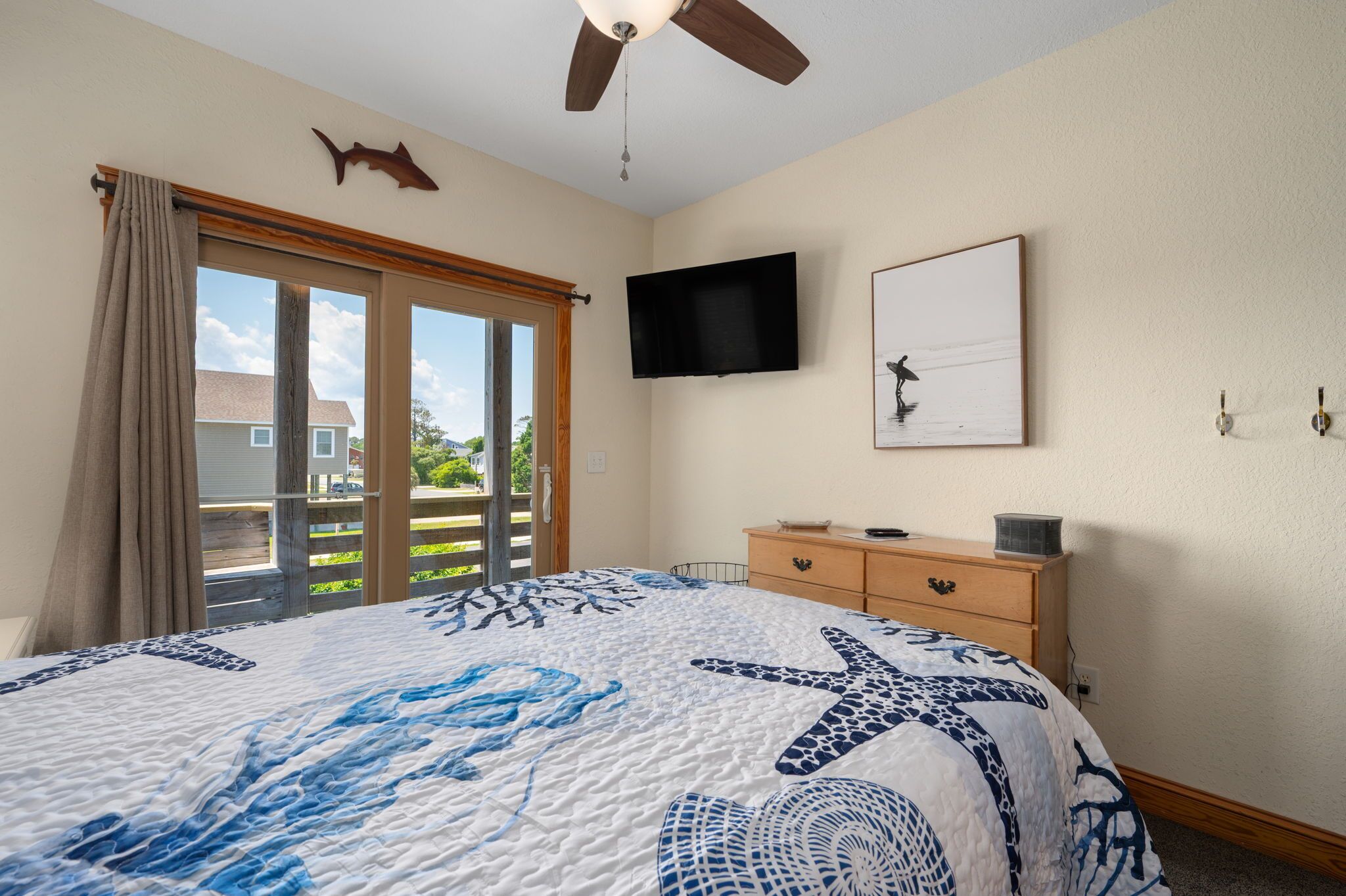 Une chambre dotée d’un grand lit, d’un ventilateur de plafond, d’un téléviseur à écran plat, d’une commode en bois et d’une porte-fenêtre coulissante donnant sur un balcon.
