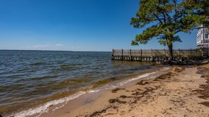 Ein Strand mit einem hölzernen Steg, der ins Wasser führt, einem Baum und einem Gebäude mit einer Umzäunung.