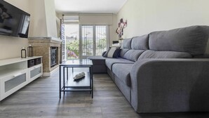 Ein modernes Wohnzimmer mit grauem Sofa, einem an der Wand montierten Flachbildfernseher, einem Kamin und einem Balkon mit Blick auf Grünflächen.