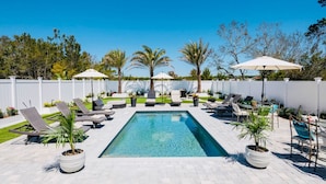 A rectangular swimming pool with lounge chairs and umbrellas, surrounded by a white fence and palm trees. -  SANDSCASTLE :Luxury: Iconic Retreat,Sleeps 16, Heated Pool, Beach Steps Away  (Panama City Beach)