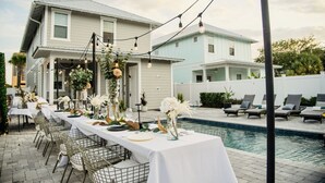A poolside dining area with tables set for a meal, surrounded by lounge chairs and a swimming pool. -  SANDSCASTLE :Luxury: Iconic Retreat,Sleeps 16, Heated Pool, Beach Steps Away  (Panama City Beach)