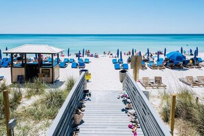 Una playa con un paseo de madera que conduce a una cabaña, sillas de playa azules y sombrillas.