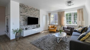 A modern living room with a flat-screen TV, a stone wall, a sofa, a coffee table, and a window with curtains. - Bright & Charming Vacation Apartment "Kille - Right Apartment" in Sankt Peter Ording (St. Peter-Ording)