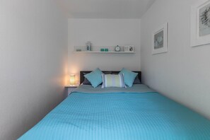 A bedroom with a bed, a nightstand with a lamp, a shelf with decorative items, and framed pictures on the wall.