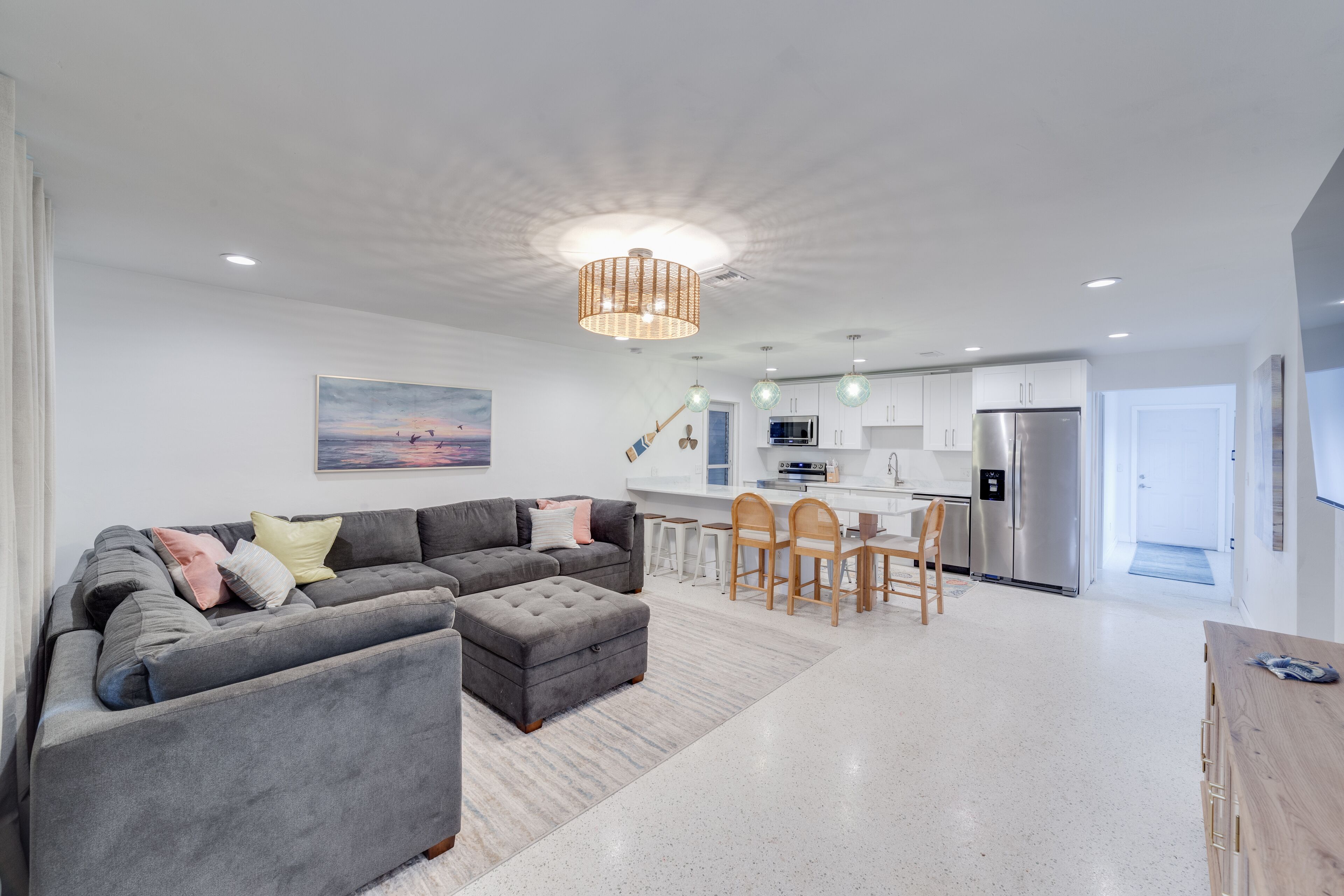 A modern living room with a grey sectional sofa, ottoman, and a dining area with wooden chairs and a table. A kitchen with white cabinets and stainless steel appliances is visible in the background.