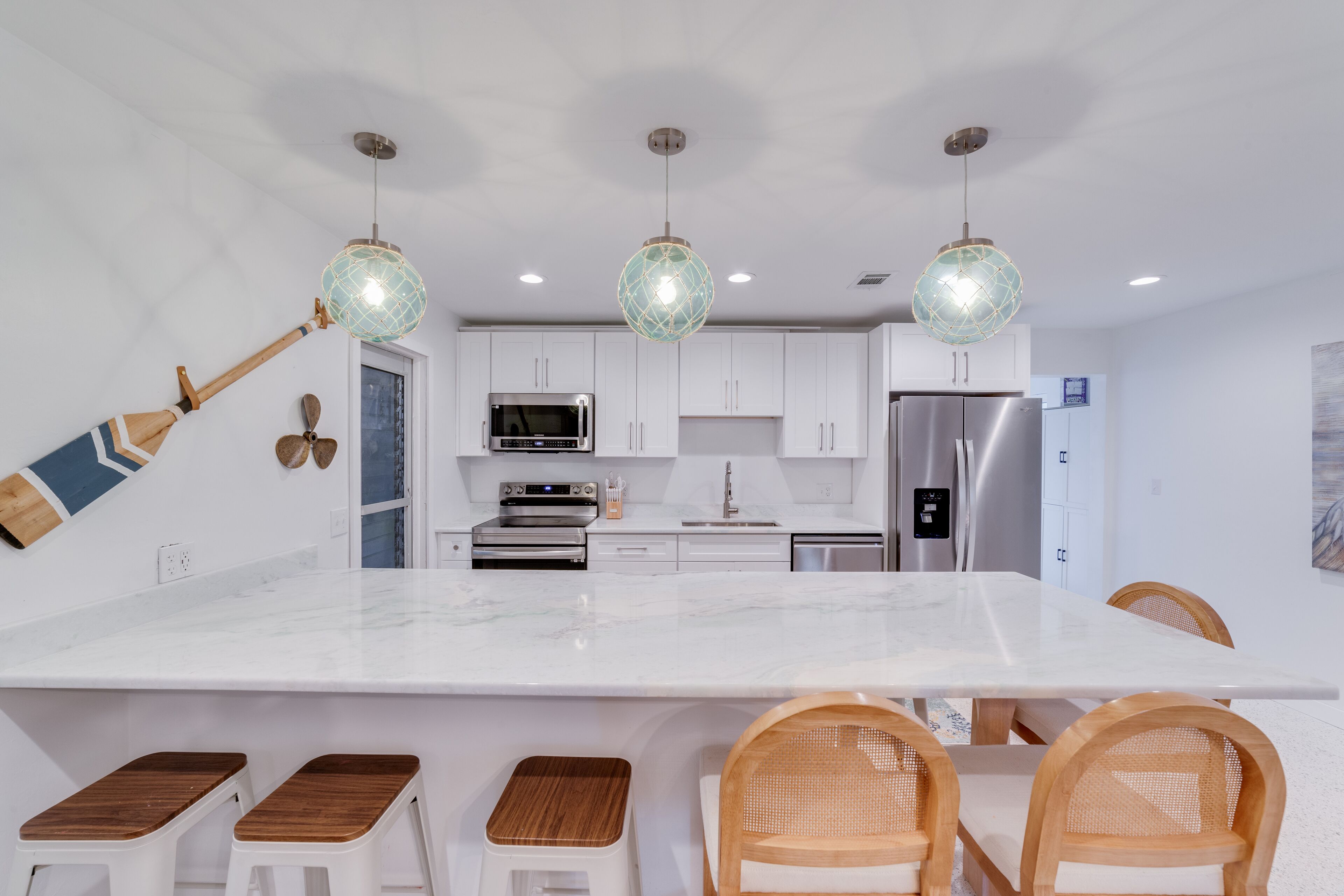 A modern kitchen with white cabinets, a large island with stools, stainless steel appliances, and decorative hanging lights.