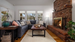 A modern living room with a brick fireplace, a grey sofa, a wooden coffee table, and a large window with blinds.