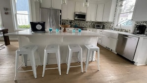 Une cuisine moderne dotée d’un îlot central, de tabourets de bar blancs, d’appareils électroménagers en acier inoxydable et de luminaires suspendus.