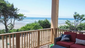Une terrasse en bois avec un canapé rouge et des coussins, donnant sur une plage bordée d’arbres et sur l’océan.