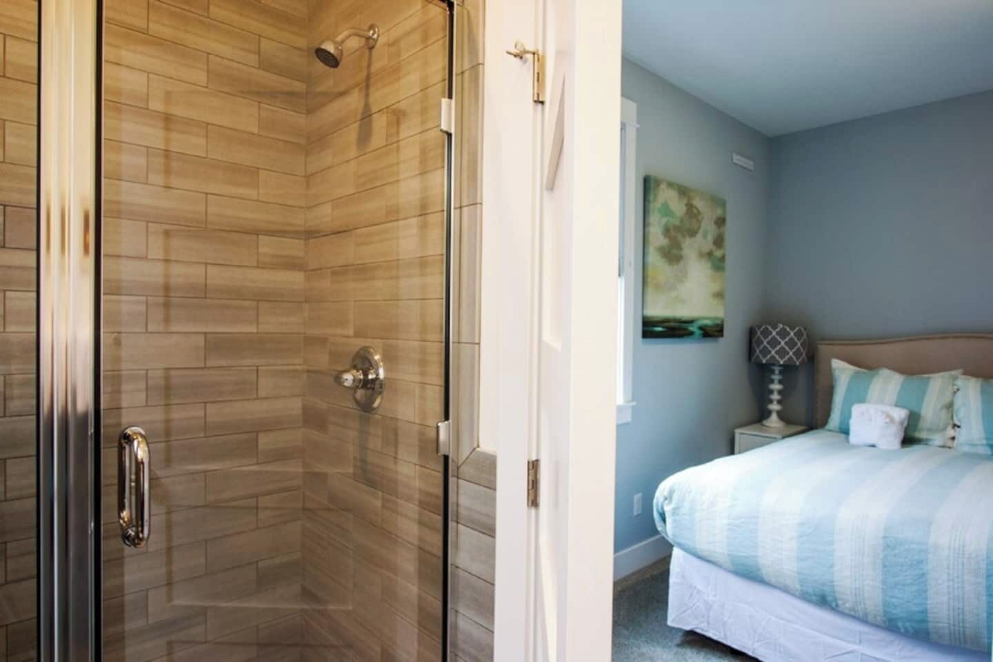A modern bathroom with a glass shower enclosure and a separate shower area. Adjacent to the bathroom is a bedroom with a bed, a nightstand, and a wall-mounted lamp.