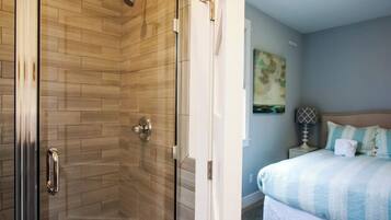 A modern bathroom with a glass shower enclosure and a separate shower area. Adjacent to the bathroom is a bedroom with a bed, a nightstand, and a wall-mounted lamp.