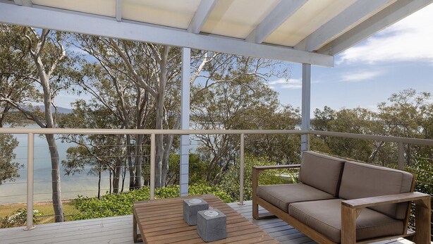 A covered outdoor area with a wooden table and a cushioned bench, overlooking a body of water and trees.