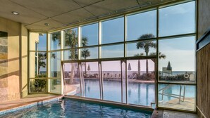 An indoor swimming pool with clear glass walls, a tiled floor, and a view of palm trees and a building outside.